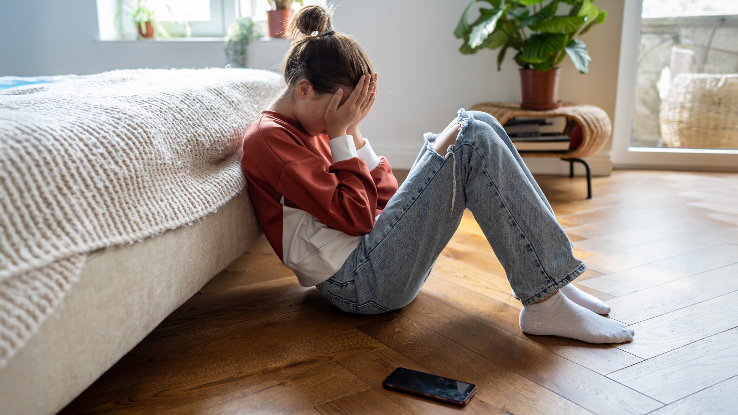Unhappy teen girl covering face with hands crying while sitting on floor with mobile phone nearby